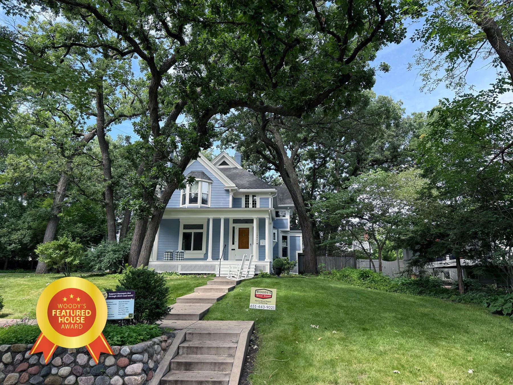 A two-story blue house with white trim and a front porch, surrounded by large trees and a well-maintained lawn, with signs in the yard indicating it is a featured house and home repair services.
