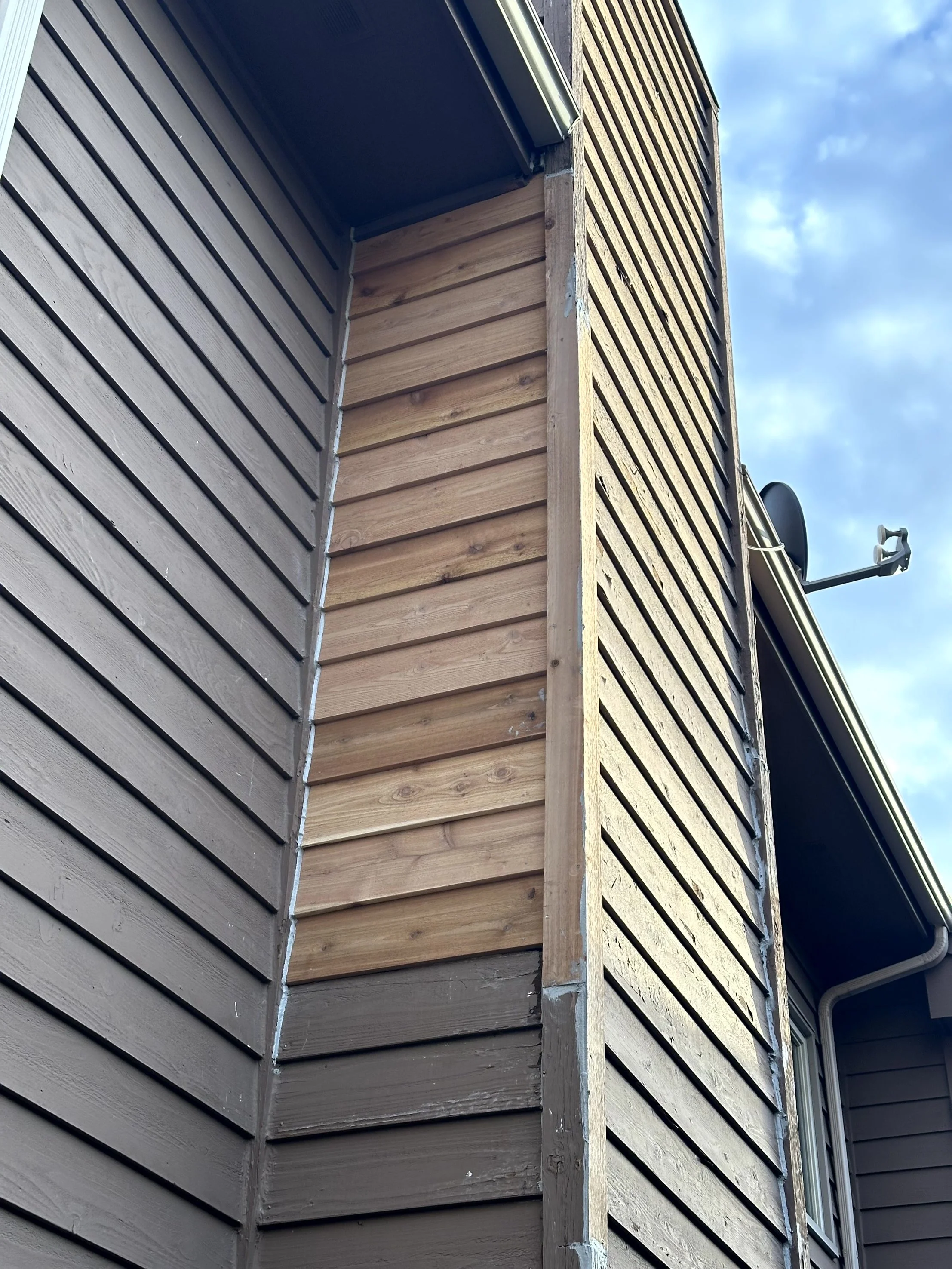 Close-up of the corner of a house showing two different siding types, one painted and one natural wood, under a blue sky with clouds.
