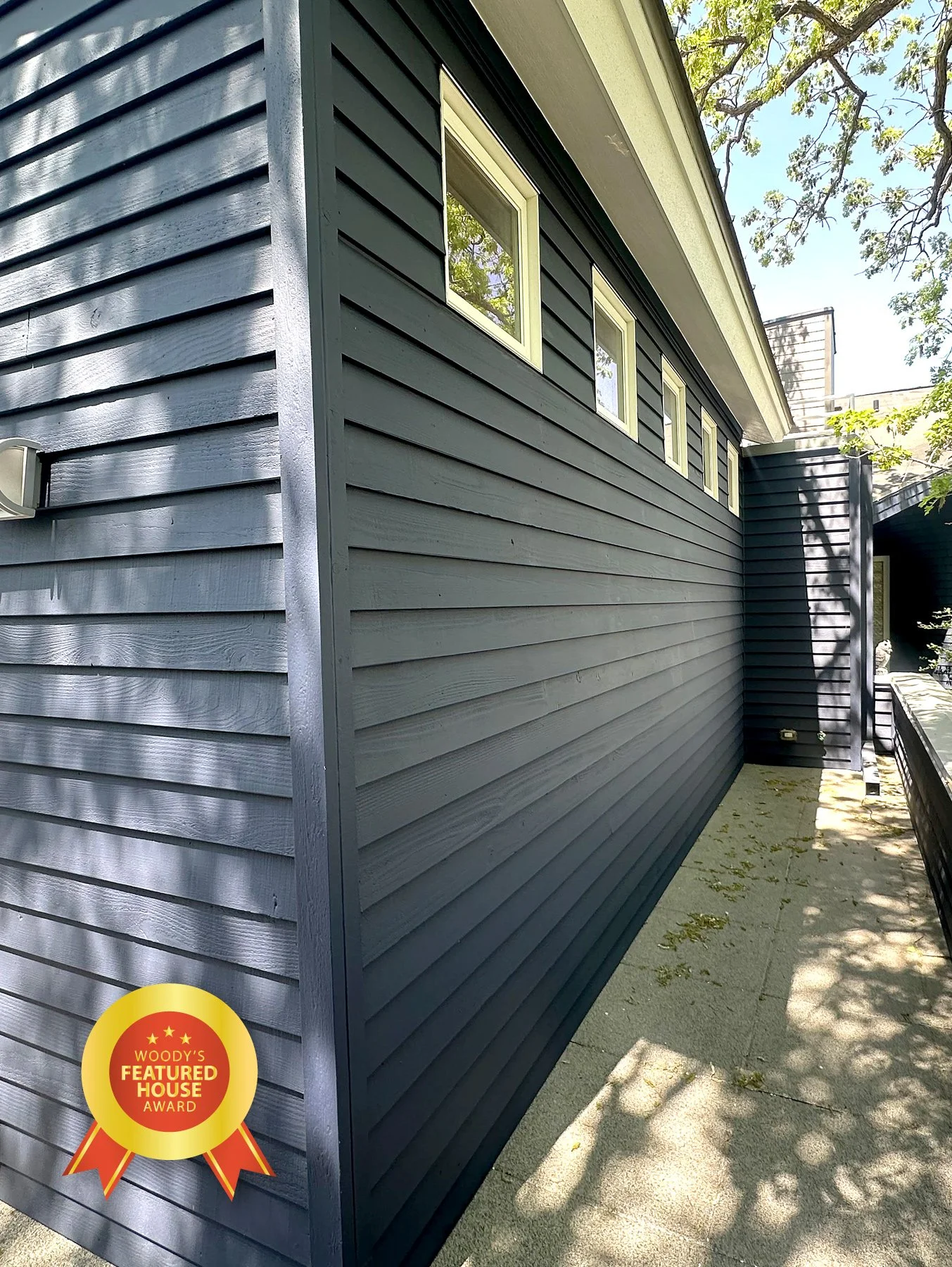 Side view of a modern house with dark gray horizontal siding, rectangular windows, and a concrete pathway, with shadows from trees on the ground. An award badge in the lower left corner reads 'WOODY'S FEATURED HOUSE AWARD.'