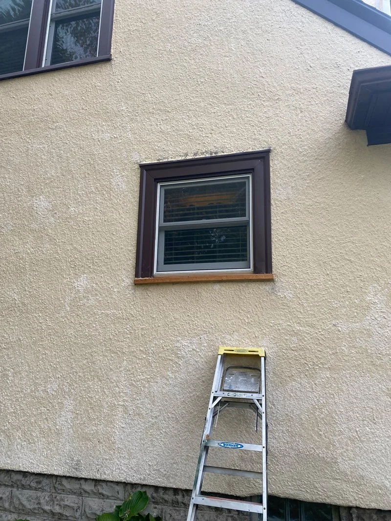 Front of a house with beige textured exterior walls, a window with dark brown trim, and a step ladder in front of the wall.