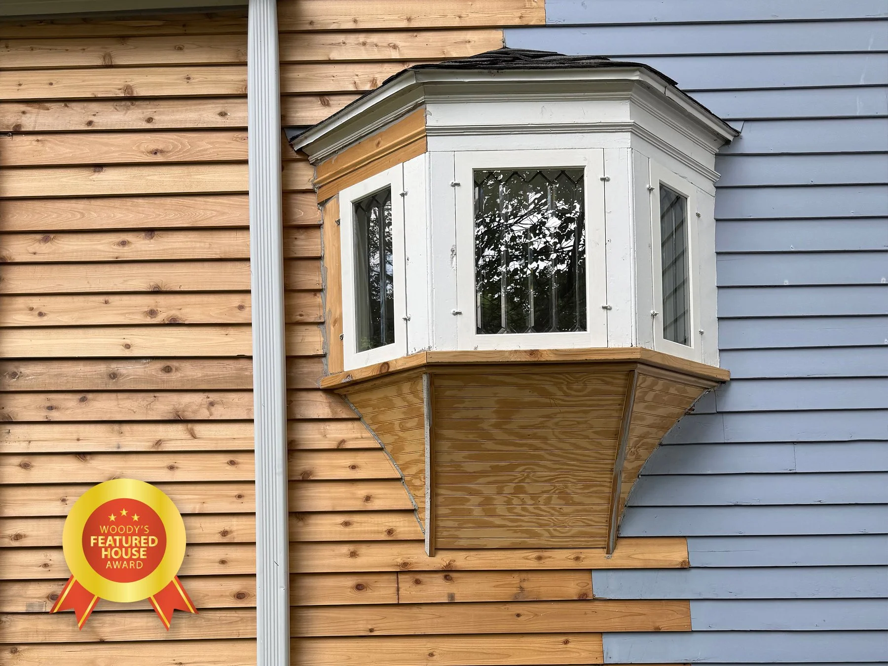 Exterior of a house with a decorative bay window. The house has split siding in natural wood on the left and painted blue on the right. There is a rain gutter and an award badge on the wall.