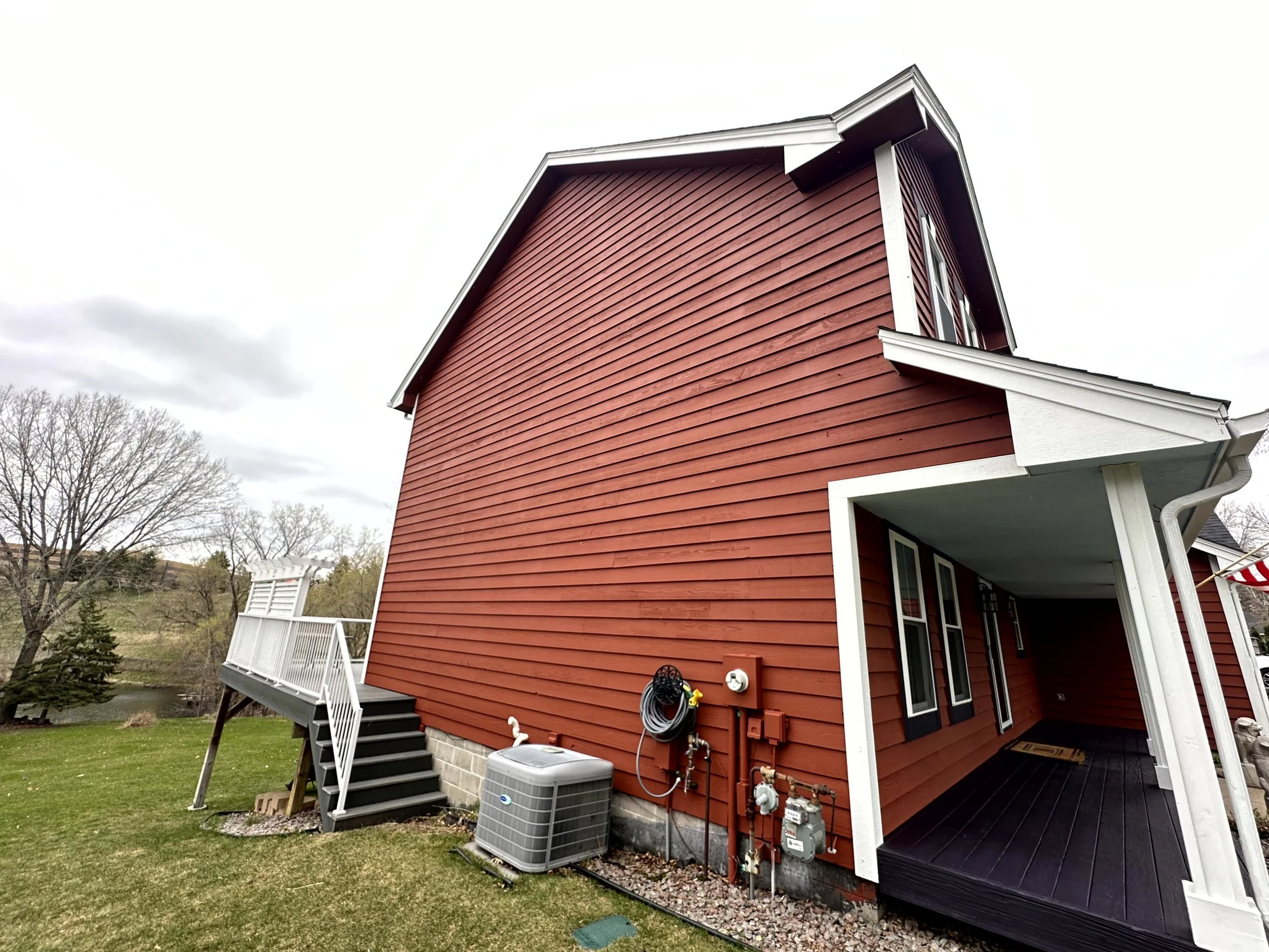 Red house with white trim, side entry porch, staircase, and outdoor HVAC unit, with a backyard and trees in the background under cloudy sky.