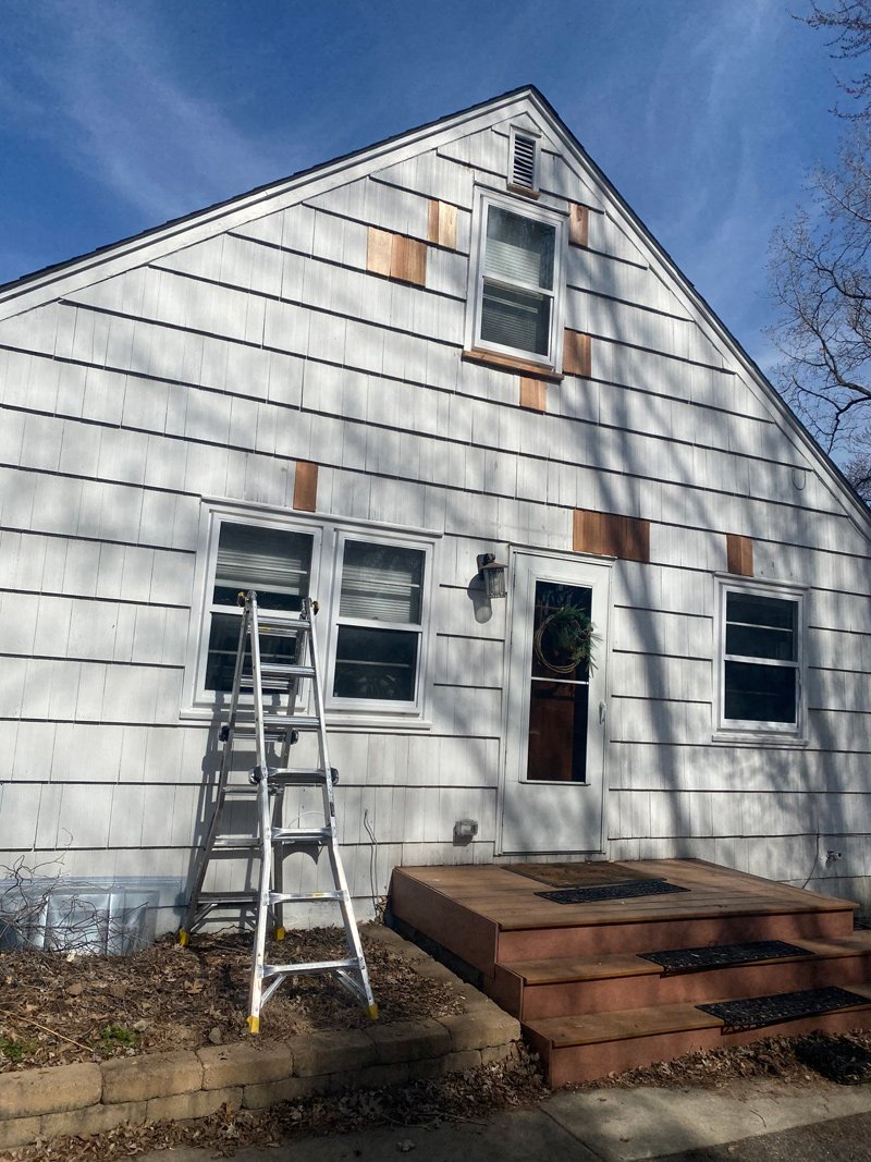 A house exterior with white horizontal siding, four windows, a porch with wooden steps, a ladder leaning against the house, and a glow to a clear blue sky.
