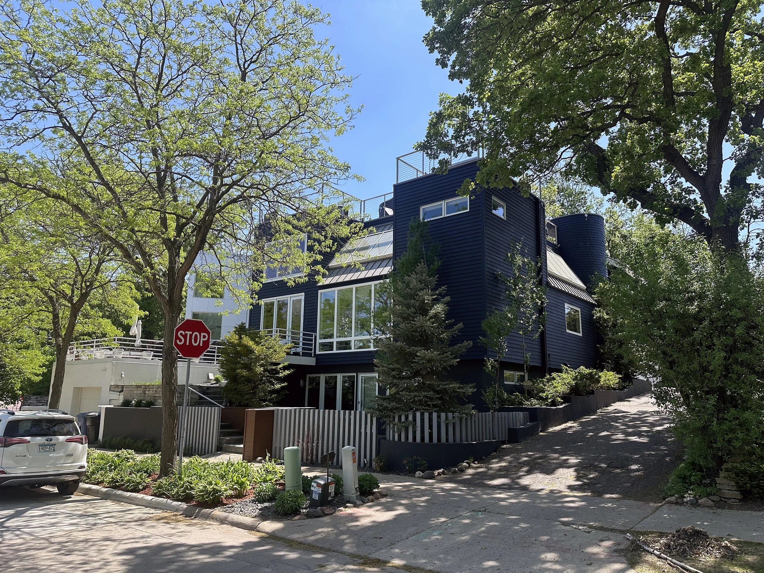 Modern blue house with large windows on a sloped driveway, surrounded by green trees and plants, with a stop sign and parked cars nearby.