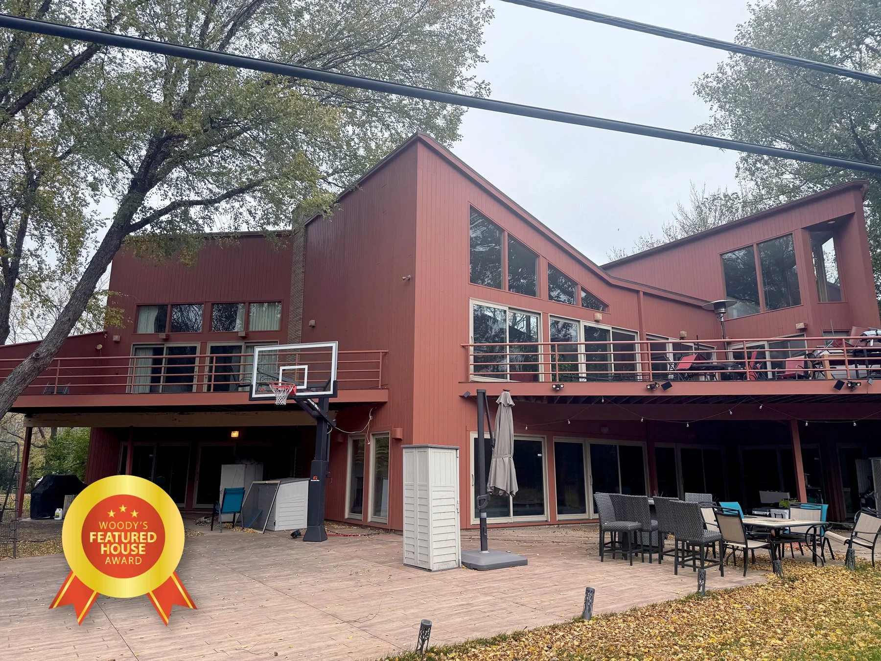 A large red modern house with multiple levels and large windows, featuring a backyard patio with outdoor furniture, a basketball hoop, and a yard. The house has a two-story structure with a deck and a lower patio area, surrounded by trees and autumn 