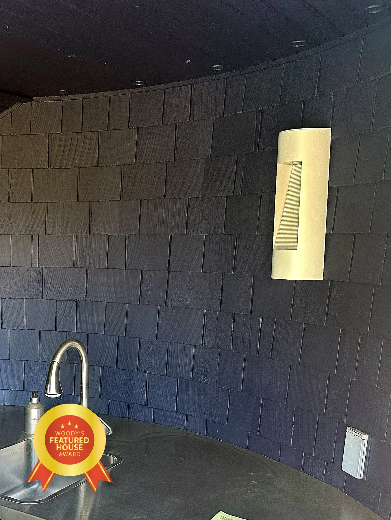 A modern kitchen counter with a stainless steel sink and a black countertop. The wall behind the counter is covered with dark gray textured tiles. There is a beige rectangular wall light fixture and a small white device or outlet on the wall. A badge
