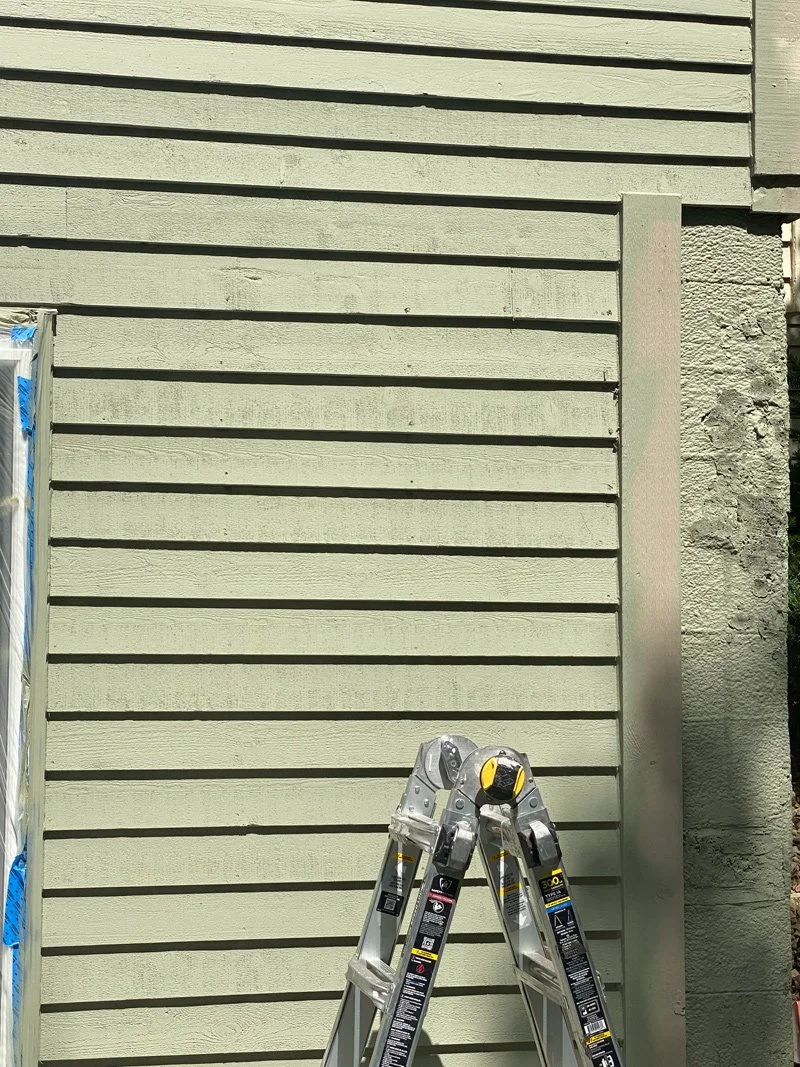 Green house siding with horizontal wooden planks, a metal support strip on the right side, and a partially visible folding ladder in the foreground.