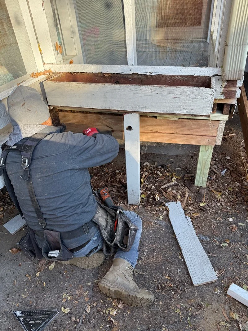 A person working on a wooden window frame outside, with some tools and wood pieces around.
