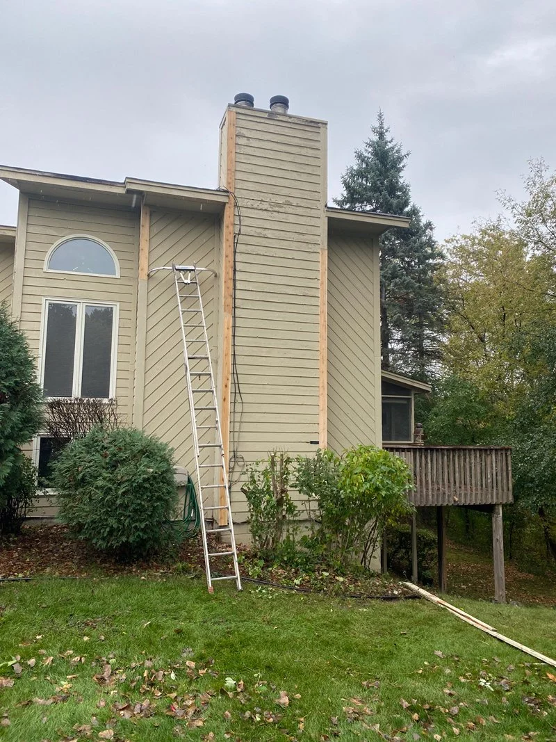 Backyard of a house with a chimney under construction, ladder leaning against the chimney, and bushes in the yard.