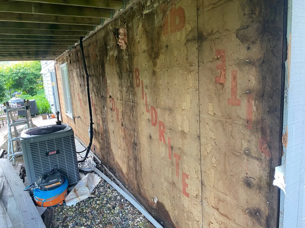 View of a house wall under renovation with plywood panels, an air conditioning unit, and construction tools, with a background of a backyard with trees and patio furniture.