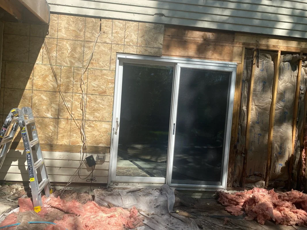 Construction site of a house exterior with a sliding glass door, insulation, exposed wood framing, plywood sheathing, and a ladder.
