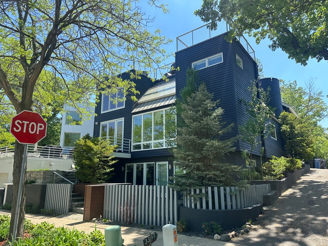 Modern blue multi-story house with large windows, surrounded by trees and greenery, with a stop sign and sidewalk in the foreground.