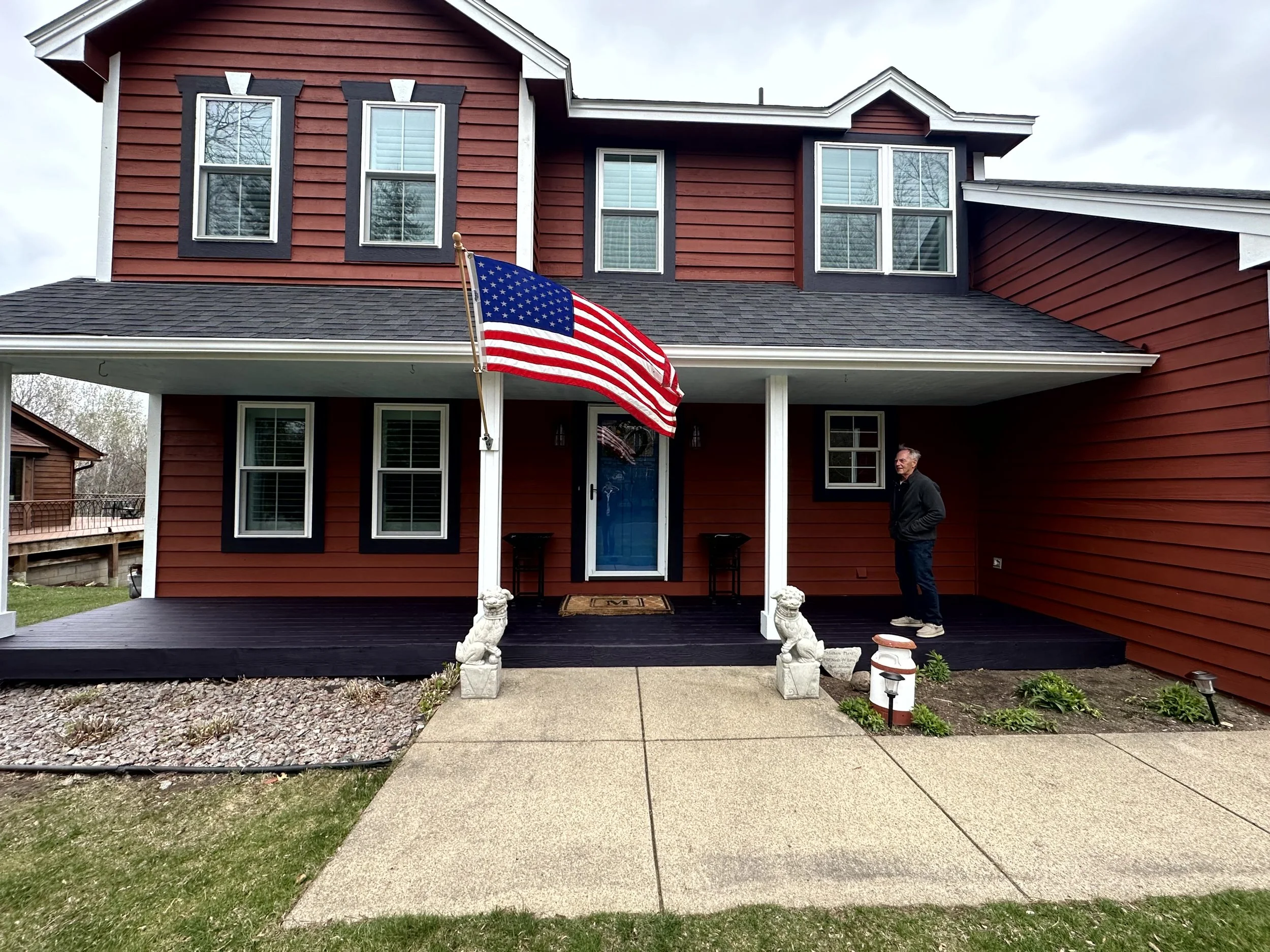 Red two-story house with black trim, American flag on front porch, man standing on porch, plants and lawn in yard.