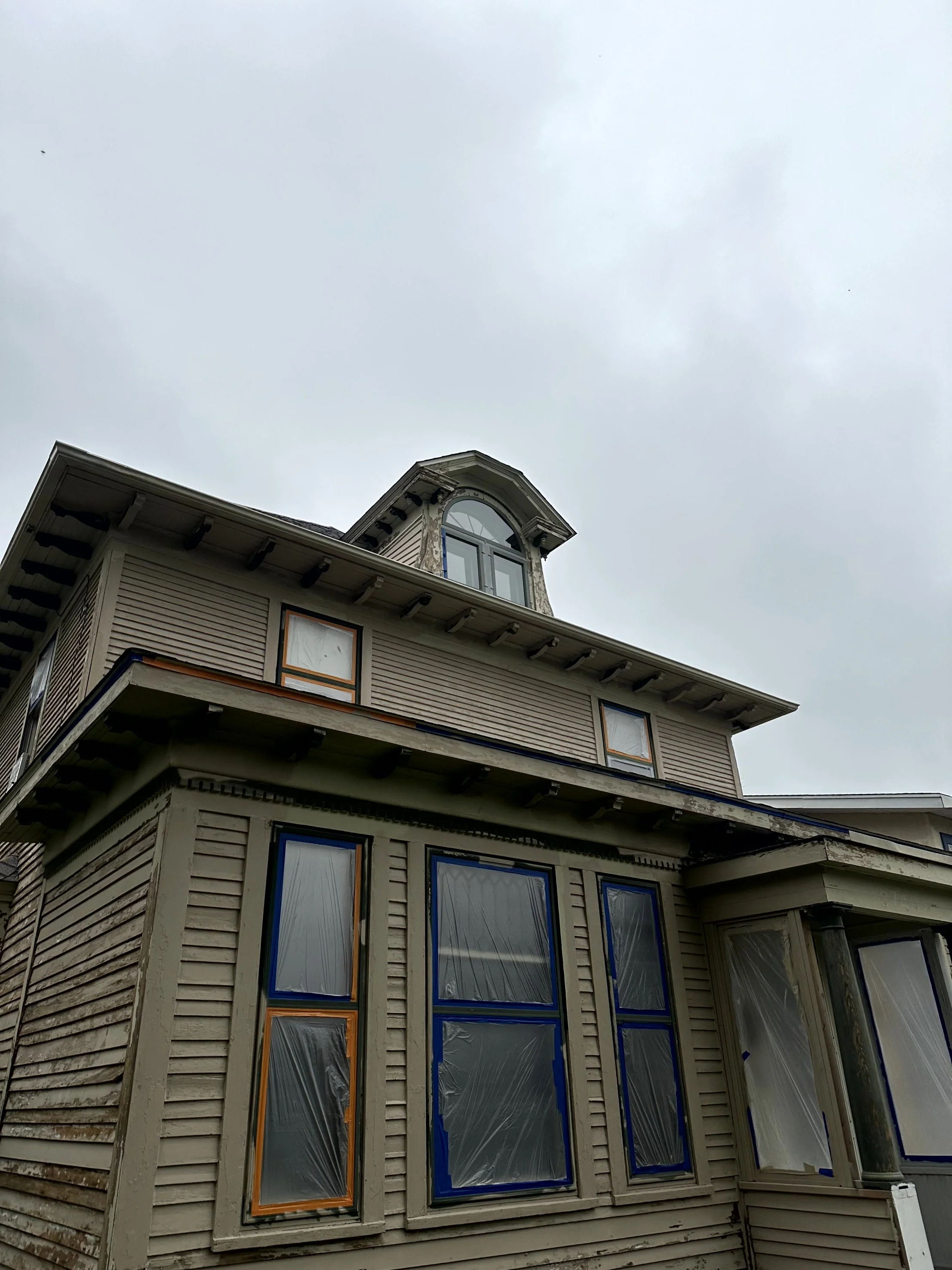 Exterior view of a multi-story house with beige siding, some windows covered with plastic, and a cloudy sky in the background.