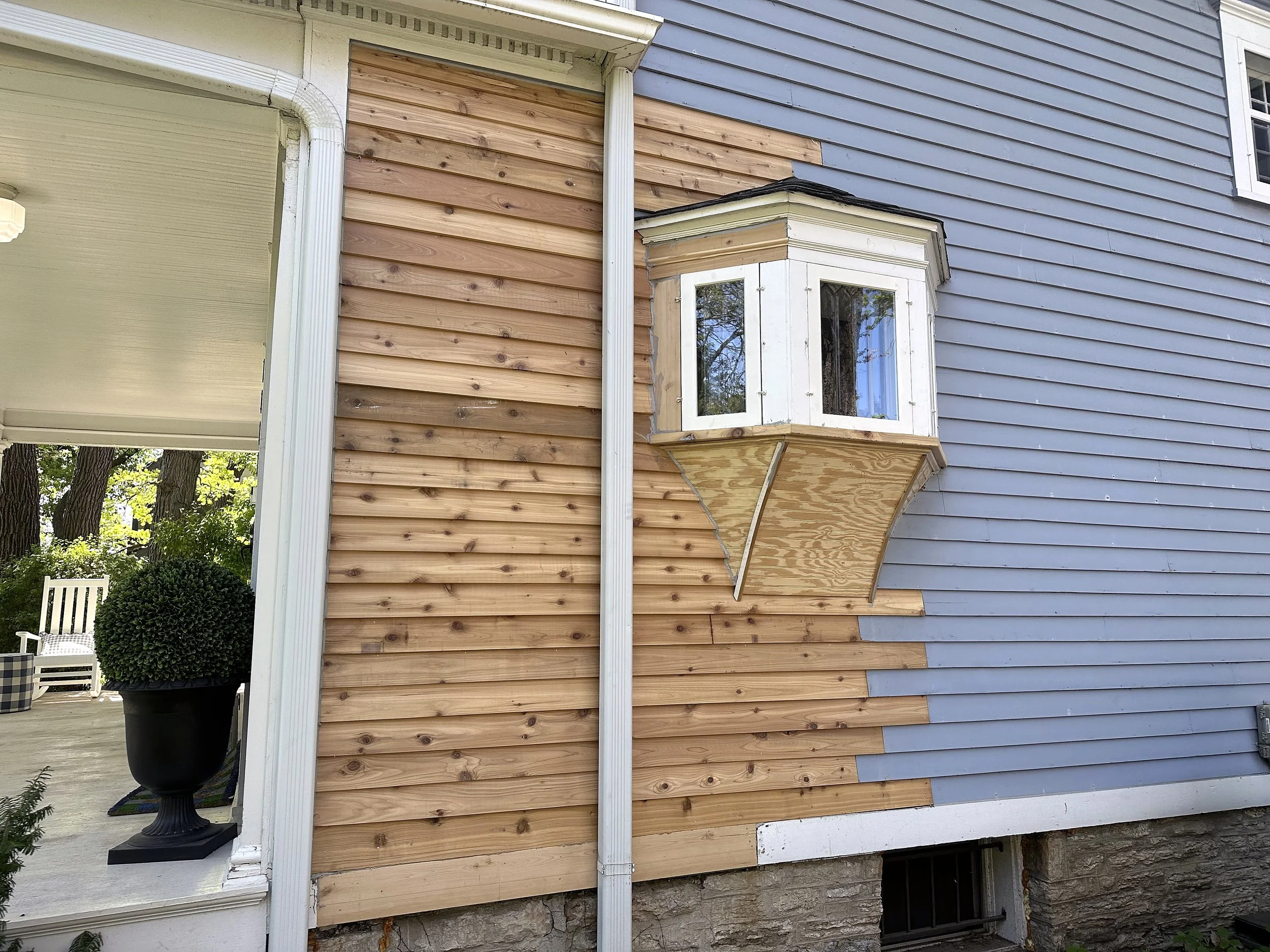 Exterior of a house with a blue painted wooden siding, an ornate white bay window, and an area of unfinished wood siding near the window.