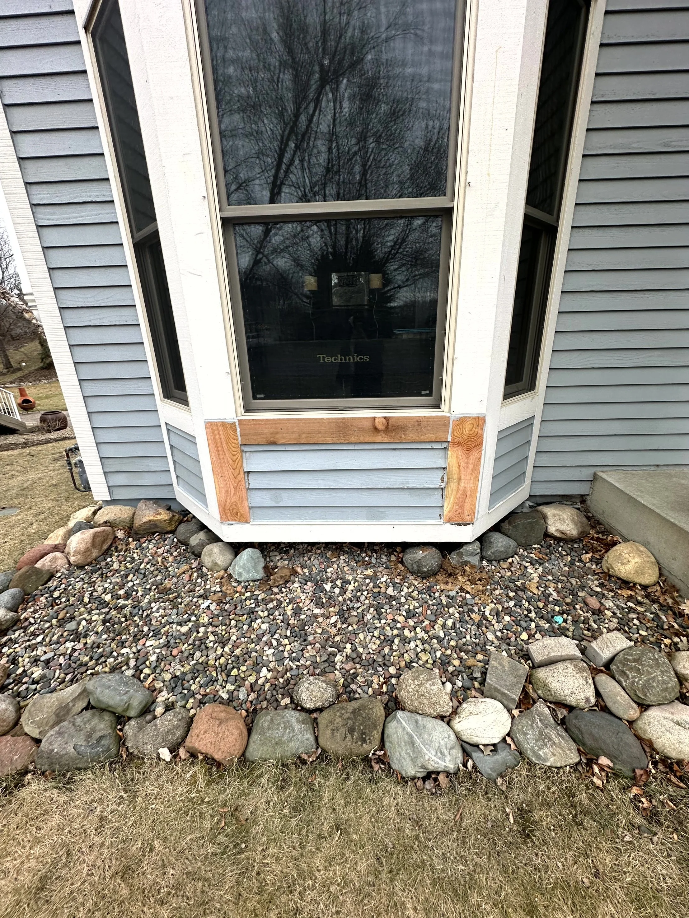Close-up of a house foundation with a decorative rock border, below a large bay window. The window has a wooden frame at the bottom, partially covered with brown wood planks. The house has gray siding and a concrete step leads to a door on the right 