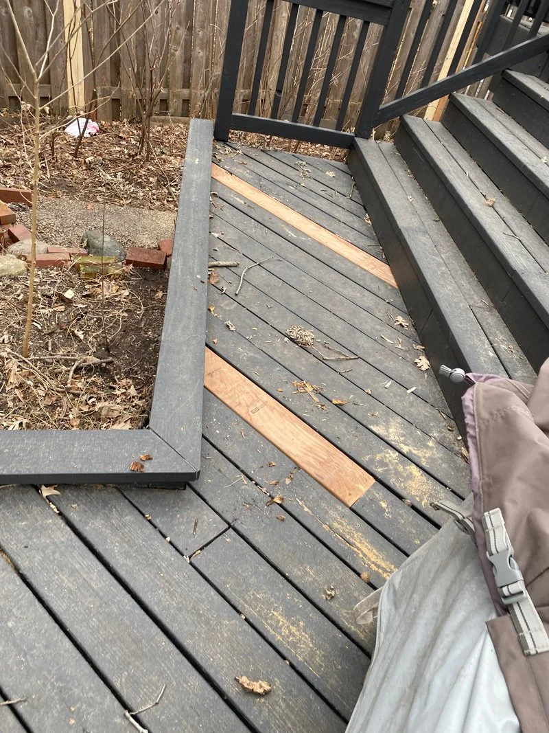 A partially repaired wooden deck with some missing middle boards, and a person using a power drill on the right side. The deck is bordered by a wooden railing and a garden area with dried leaves and small plants.