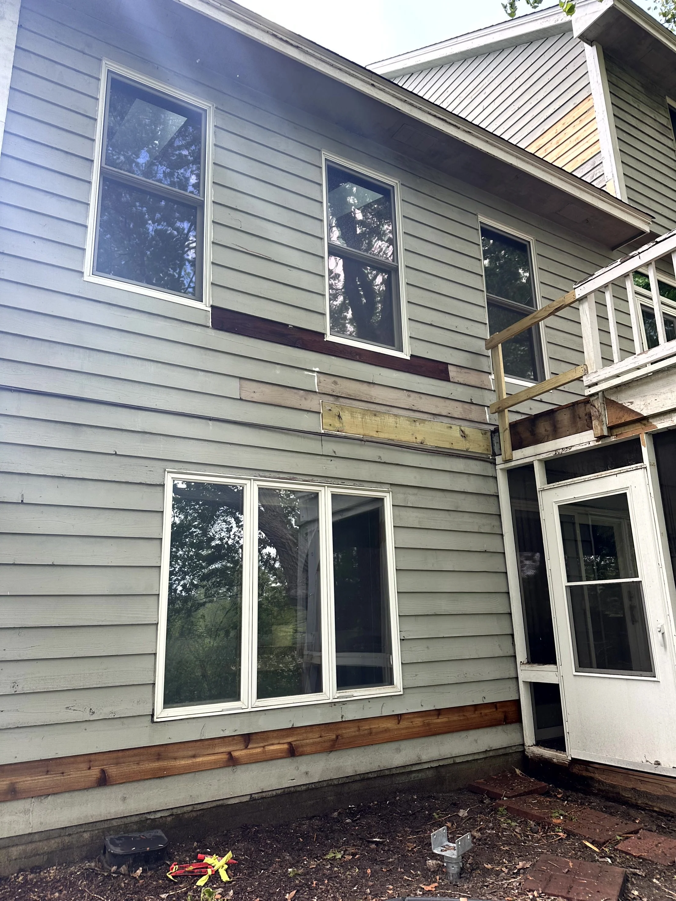 Back of a house with light green siding, multiple windows, and a small balcony under construction with unfinished wood railings. The yard has dirt, some bricks, and construction tools.