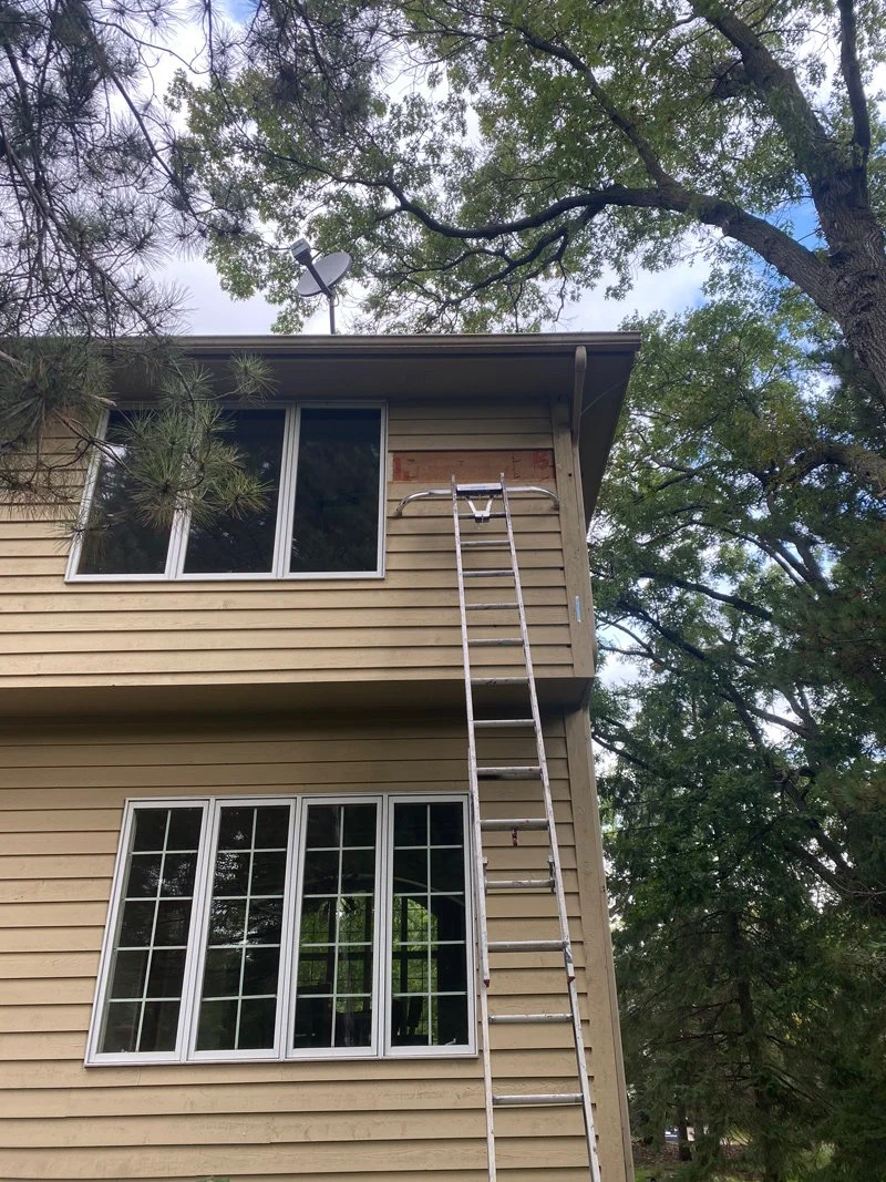 A two-story house with beige siding, two large windows on the lower floor, and a sliding glass door with a window above it on the upper floor. An extension ladder is leaning against the upper part of the house, reaching near a small section of missin
