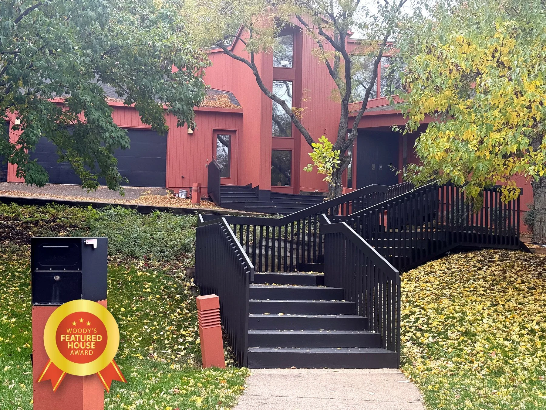 Red house with large glass windows, stairs with black railing leading to the entrance, trees with green and yellow leaves surrounding the house, fallen leaves on the ground, and a mailbox with an award sticker in the front yard.