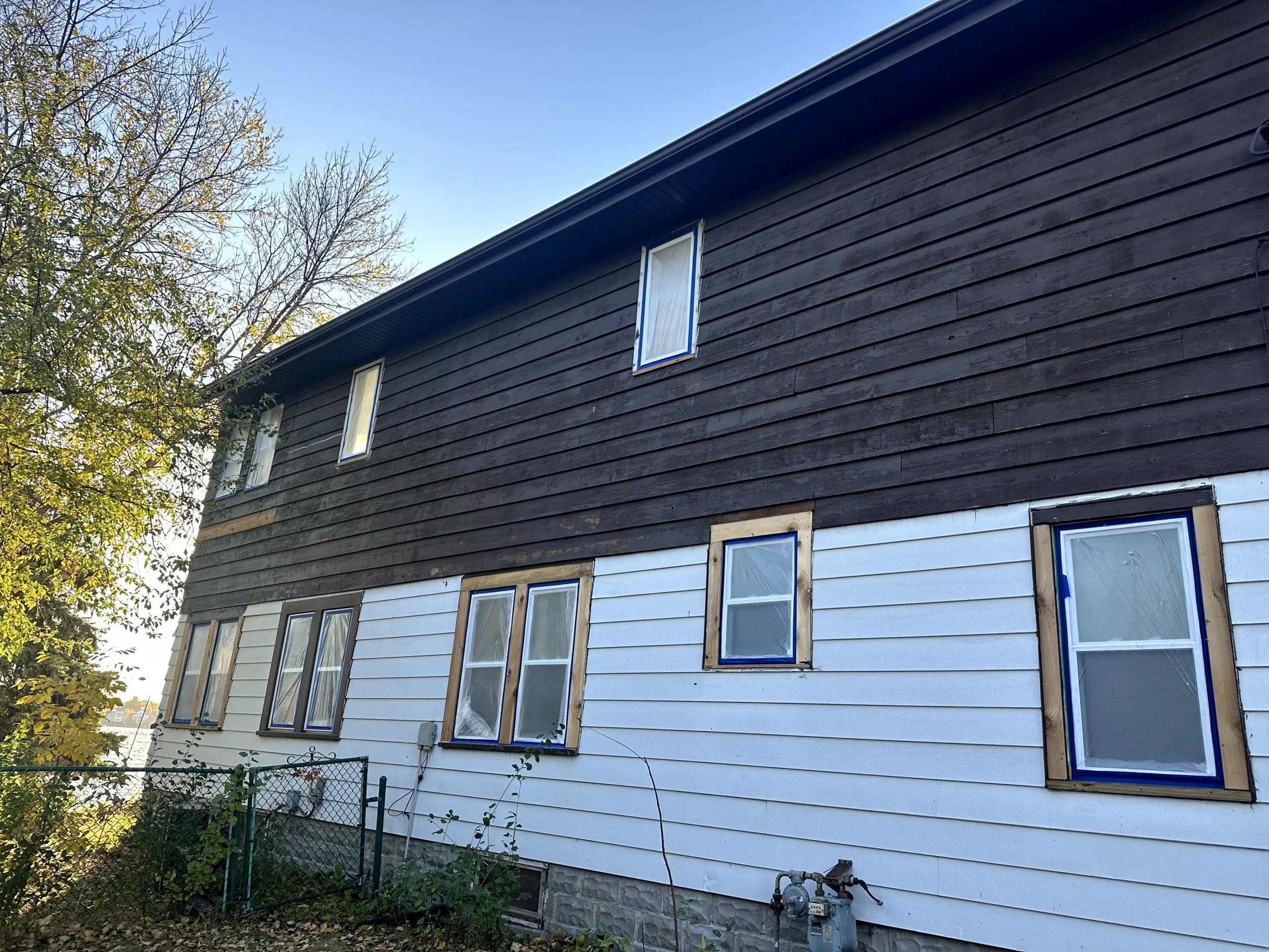 Side of a two-story house with black upper siding and white lower siding, and six windows with blue painter's tape around the frames, under a clear blue sky with some trees nearby.