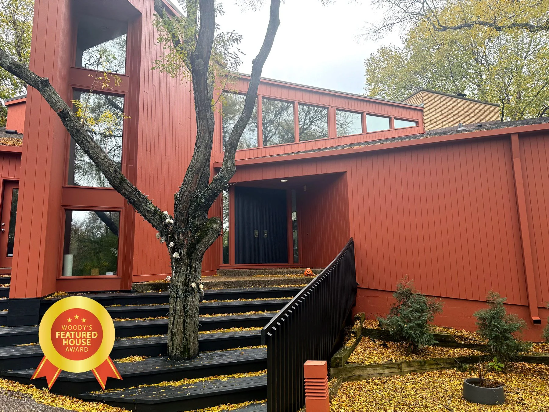 A modern red wooden house with large windows, a black front door, and a small pumpkin on the stairs. A tree with some white decorations is in front of the house. Yellow leaves cover the ground. A round award badge on the bottom left corner reads 'Woody's Featured House Award'.