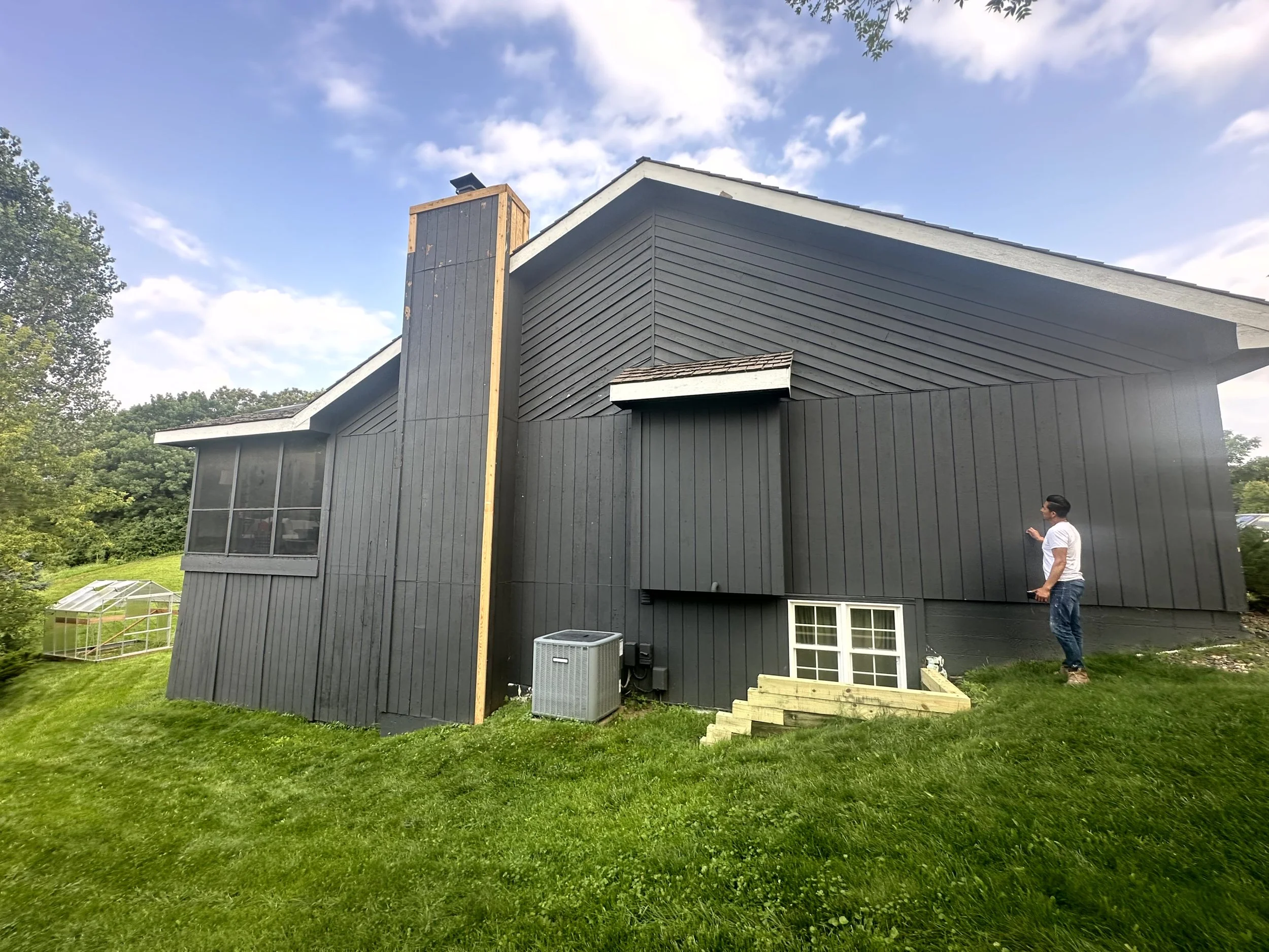 A man standing on a small set of wooden steps outside a dark gray house, with a large chimney on the left side, on a grassy yard under a partly cloudy sky.