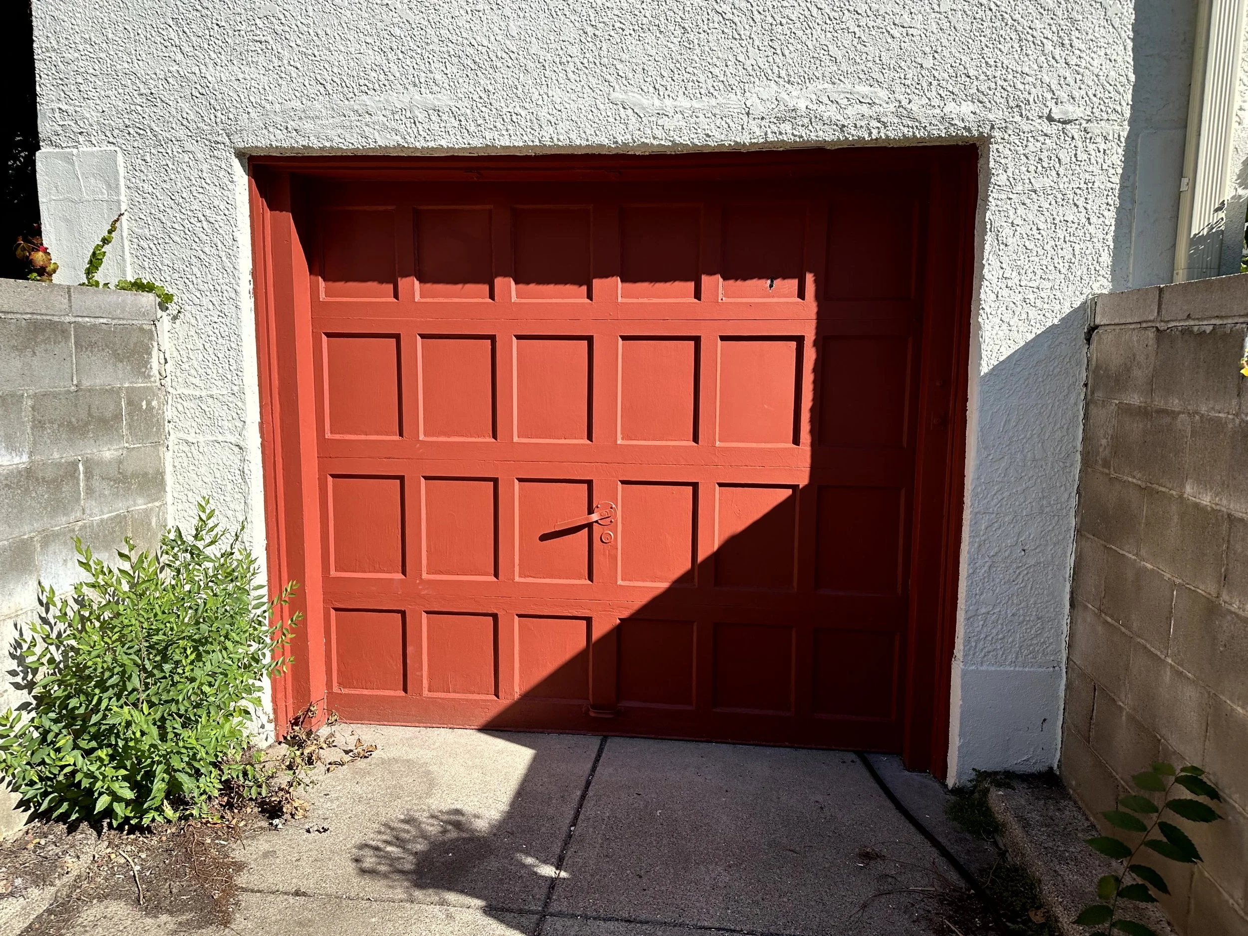 Red garage door against a white textured wall, with plants and concrete on either side and a cast shadow.