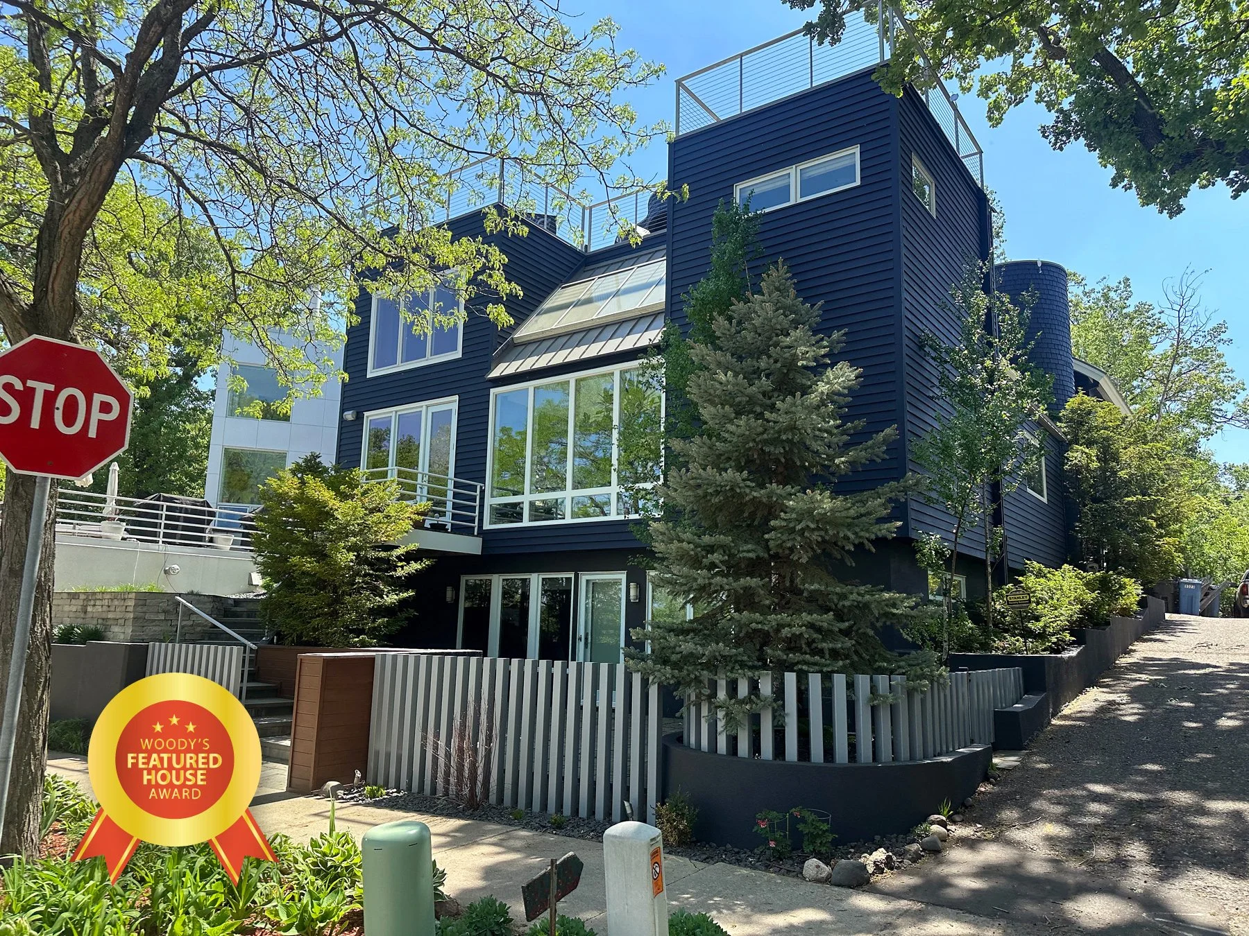 Modern multi-story house with dark blue exterior siding, large glass windows, and rooftop terraces, surrounded by trees and greenery in a residential neighborhood.