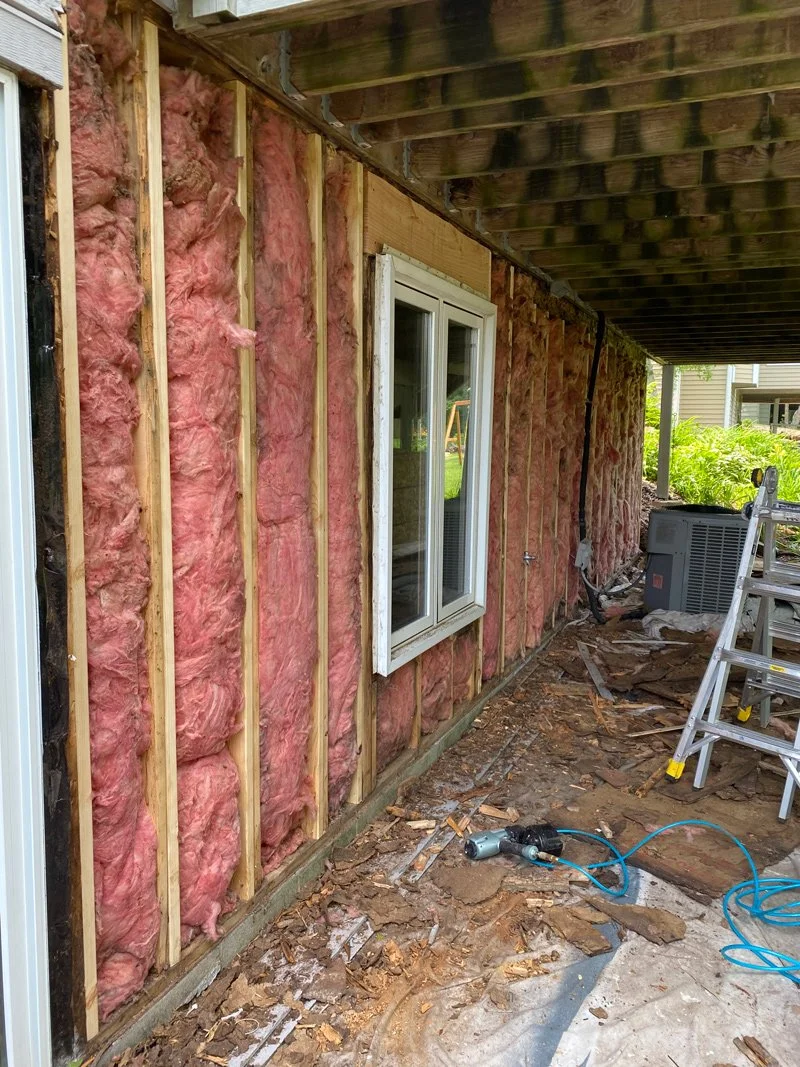 Exterior wall undergoing renovation with pink insulation and wooden framing. A window installed with white trim. Construction tools and debris on the ground, with a ladder and an HVAC unit nearby.