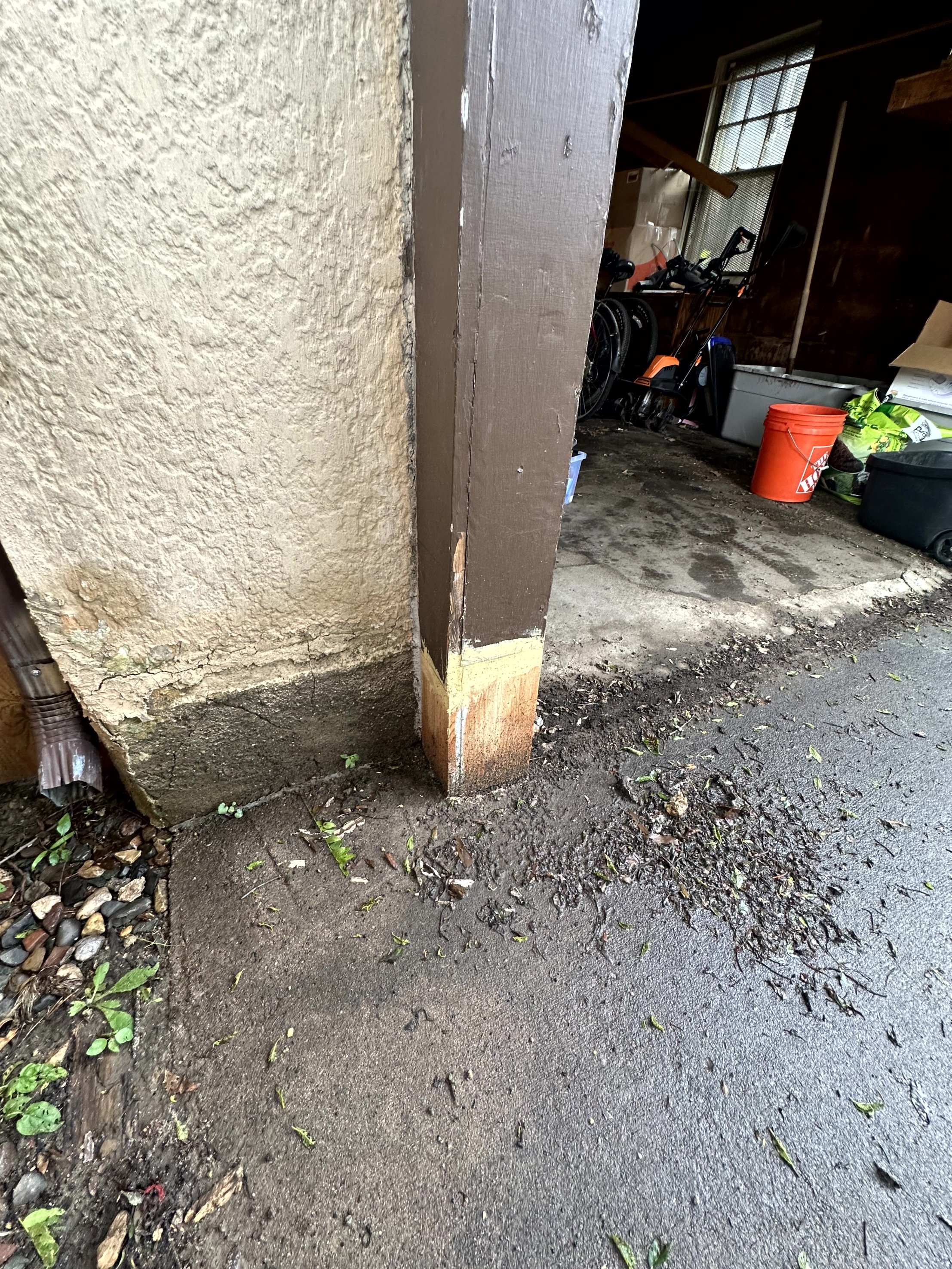 Close-up of the bottom part of a wooden garage post with exposed wood at the base, next to a textured wall and dirty concrete ground with small plants and twigs.