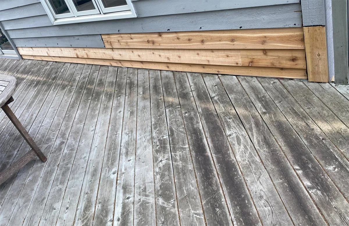 A wooden porch with weathered gray planks, part of a house with gray siding, some new wooden siding being installed, and a small section of a window visible.
