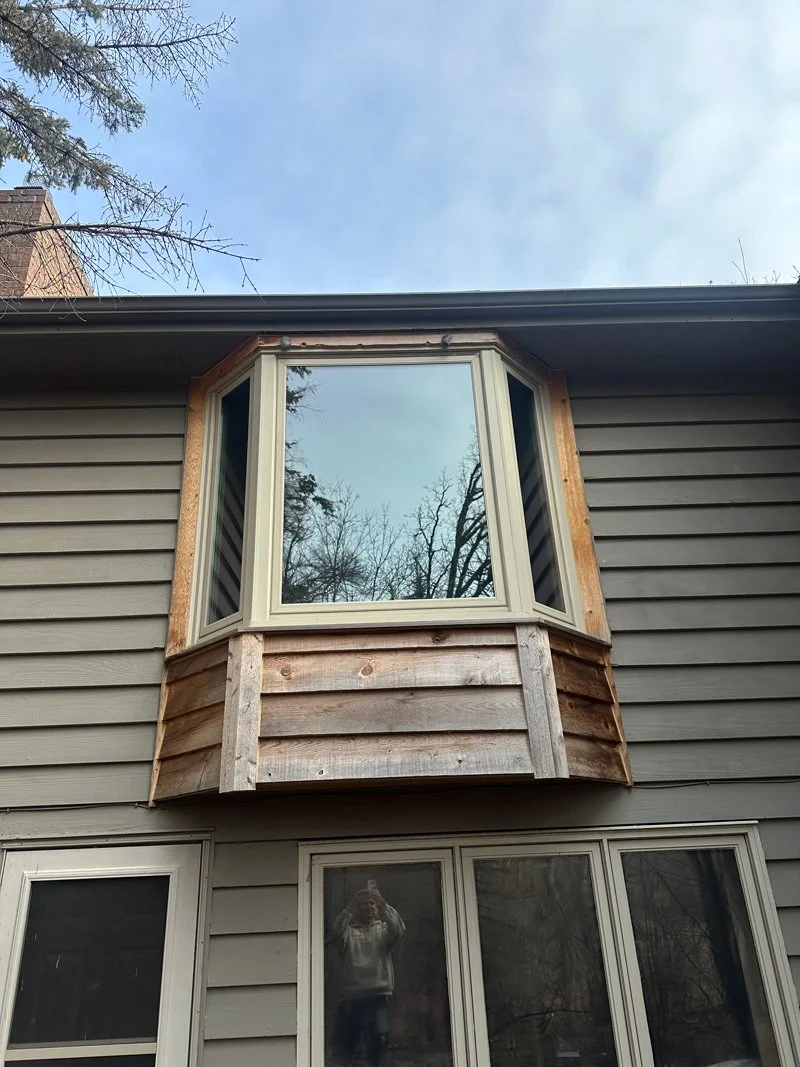 Newly installed bay window in a house's upper floor with unfinished wood framing, surrounded by gray vinyl siding, and old windows below, with trees and blue sky reflected in the glass.