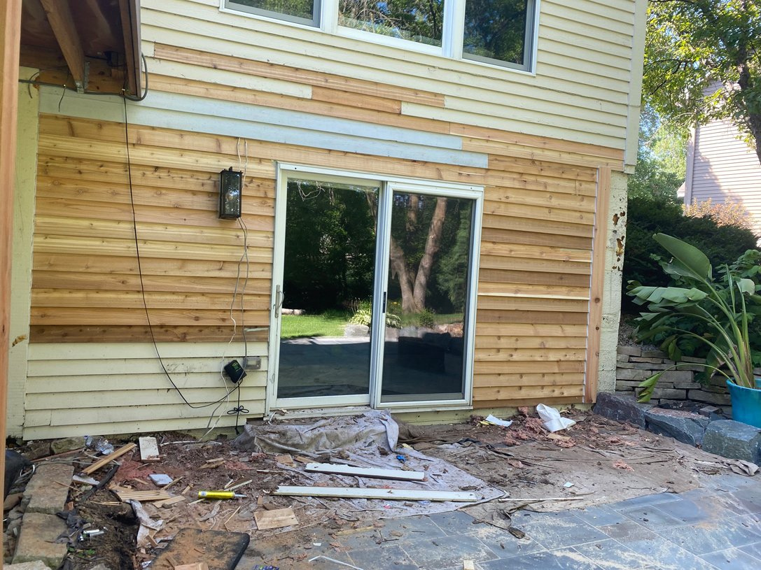 Construction site outside of a house with sliding glass door, partially installed siding, and construction debris on the ground.