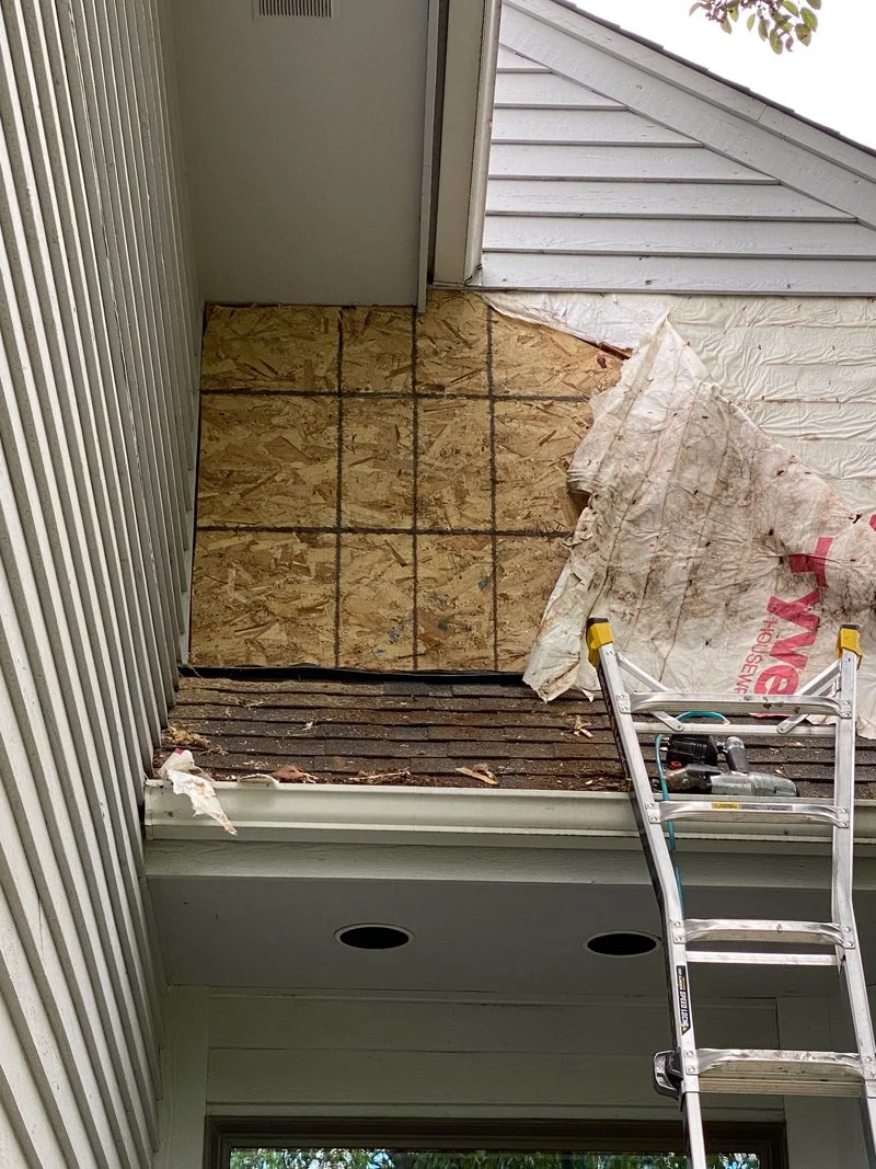 A small balcony or porch area under construction or repair, with exposed plywood flooring marked with grid lines, a metal ladder, and partial building siding and roof visible.