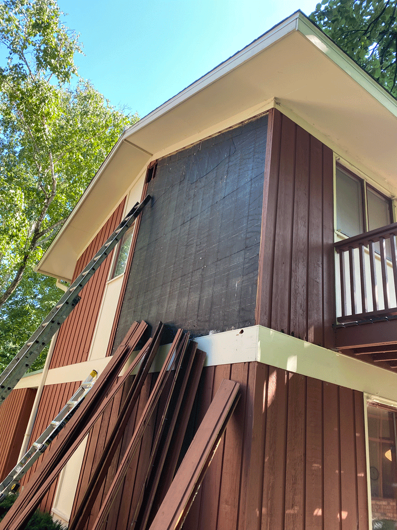 The image shows the exterior of a two-story house with a section of the siding being replaced or repaired. A ladder leans against the house, reaching up to where workers might be working. Several pieces of wood siding are stacked on the ground, and t