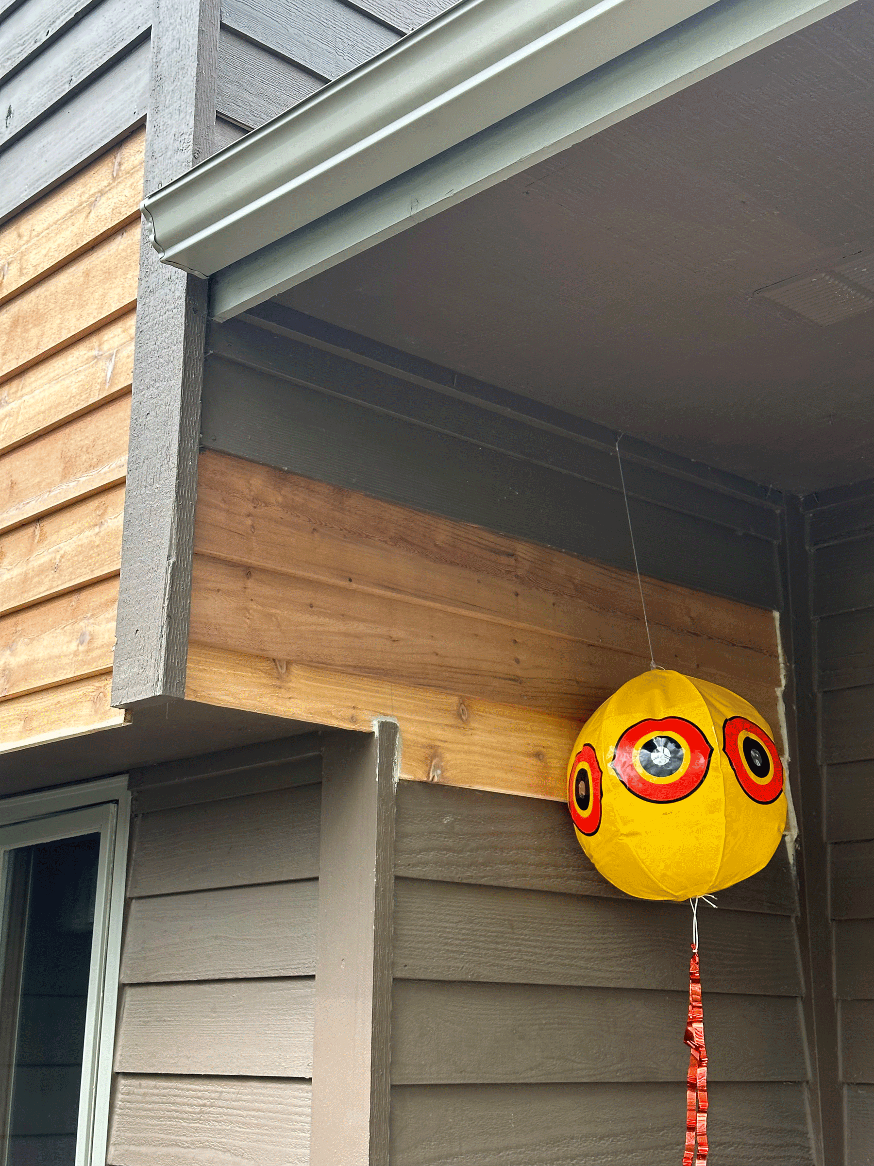 Yellow paper lantern decorated with red and black circles hanging outside of a house with gray siding and wood paneling.