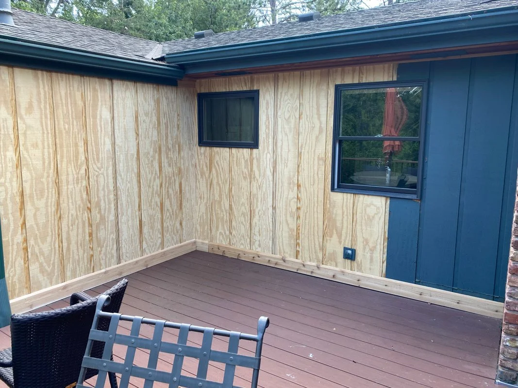 Small outdoor patio area with new wooden fencing and decking, two black and grey chairs, surrounded by a house with tan and blue exterior walls, windows, and a brick section.