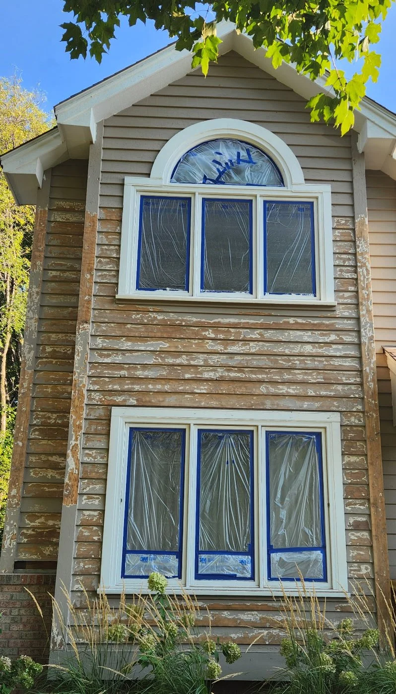 Side of a two-story house under renovation with peeling brownish-gray siding, white framed windows covered with protective plastic, and green foliage at the top.