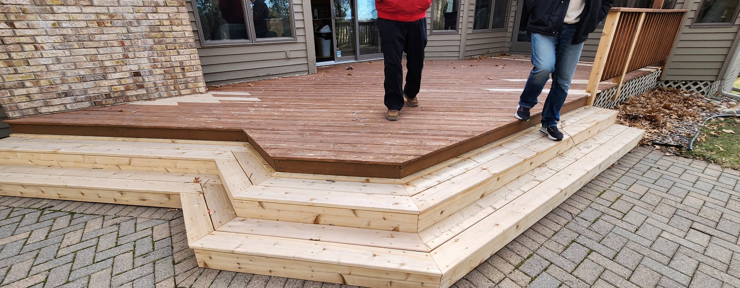 People installing a wooden deck on a patio outside a house with brick and siding exterior.
