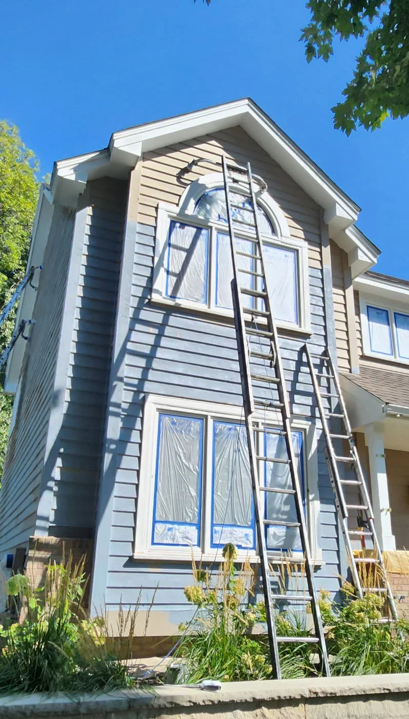 A house with light gray siding under renovation or painting, with plastic coverings on the windows, a tall ladder leaning against the house, and a bright blue sky with some green tree leaves visible at the top.