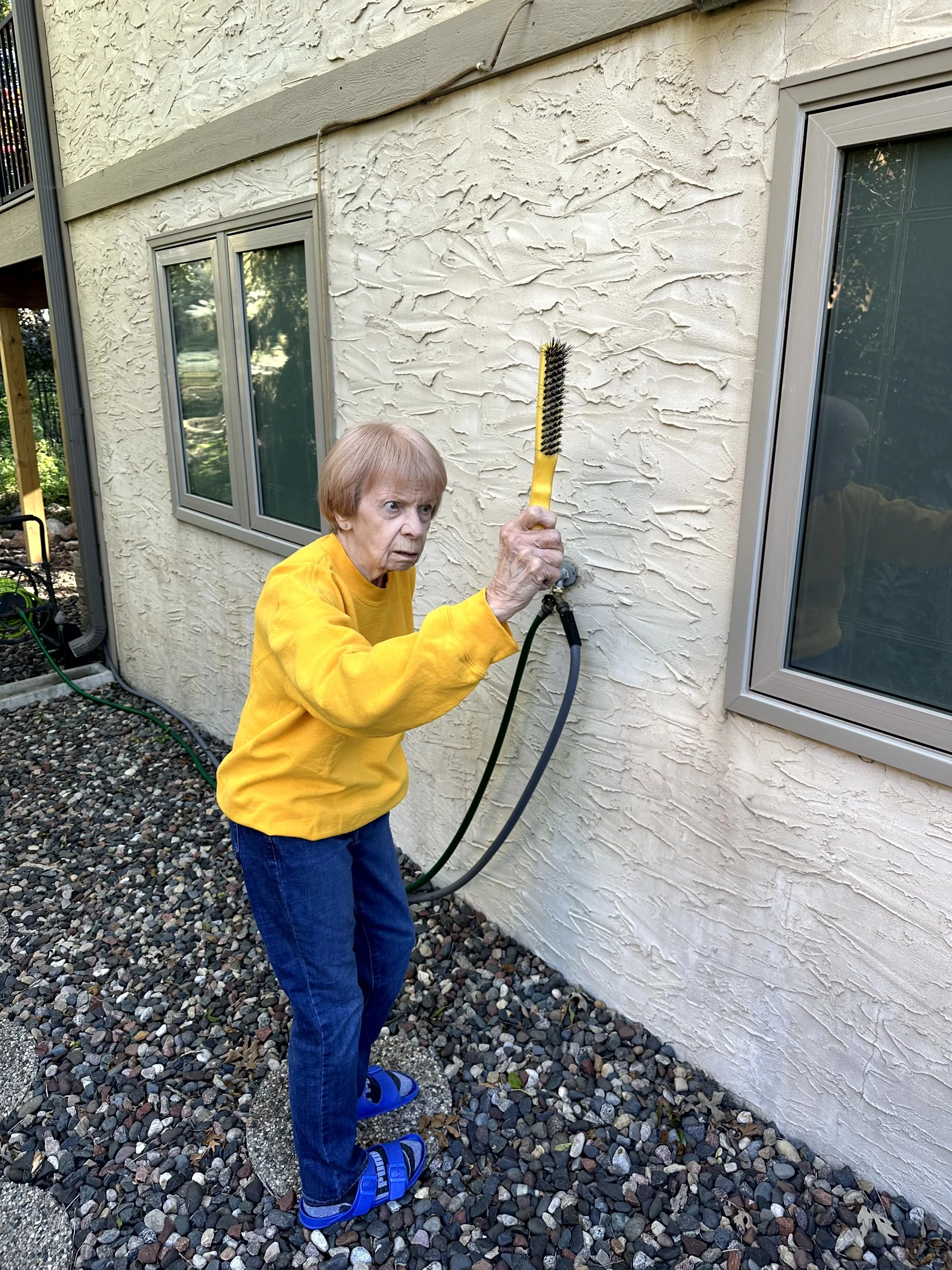 An elderly woman wearing a yellow sweatshirt, blue jeans, and blue sandals, holding a brush connected to a garden hose, cleaning the exterior wall of a house with textured beige stucco.