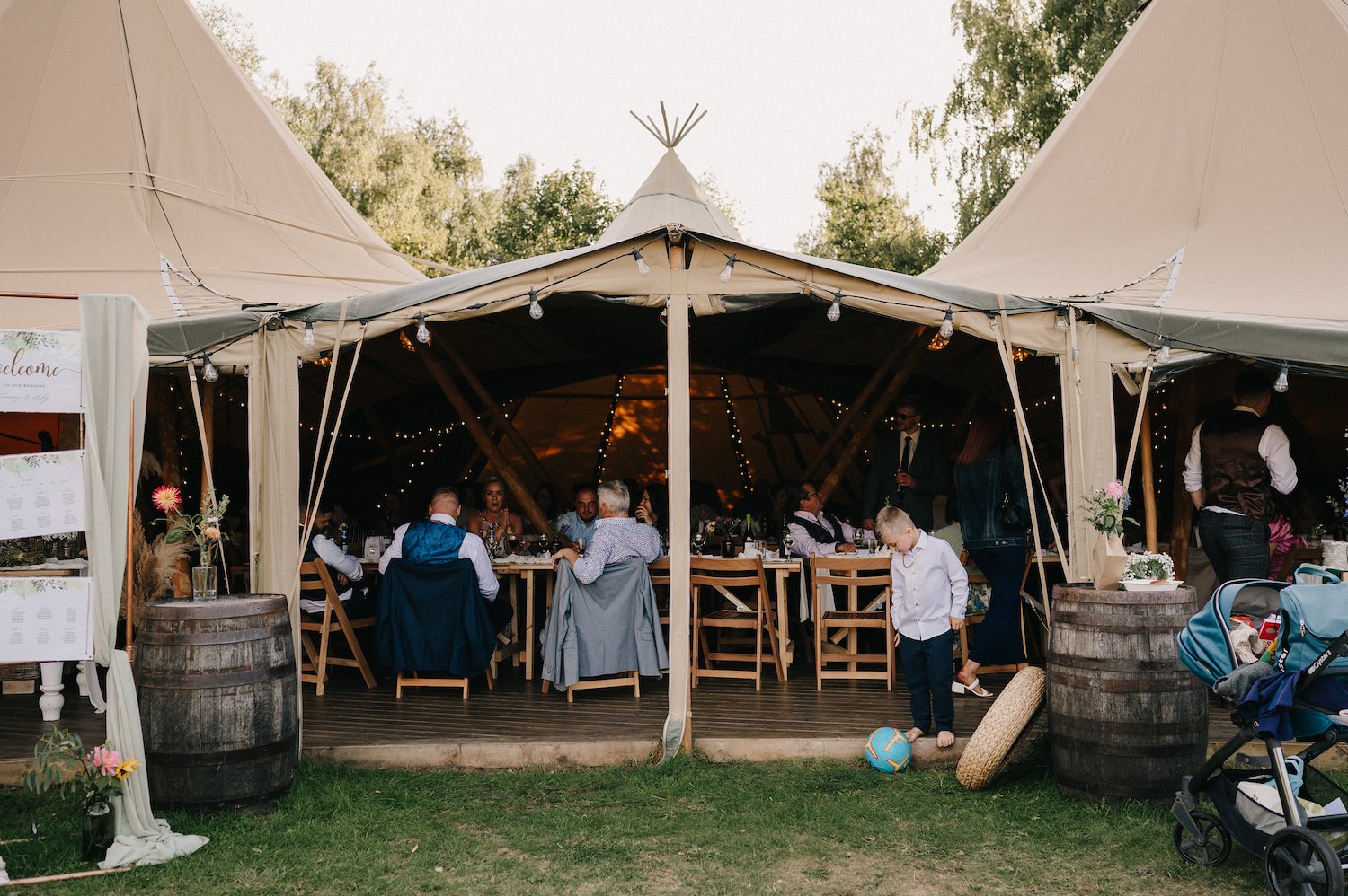 Guests sitting and standing inside a large beige tent at a wedding reception, with some children playing outside near a soccer ball on the grass and a stroller nearby.