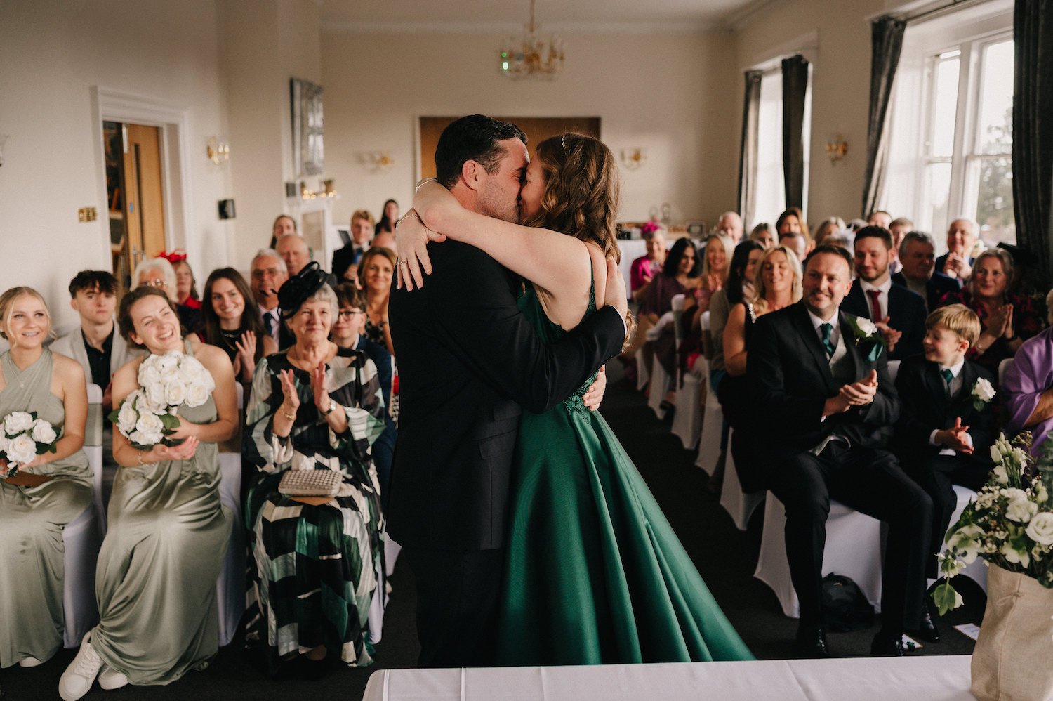 A couple kisses during their wedding ceremony in a decorated indoor venue, with guests smiling and clapping in the background.