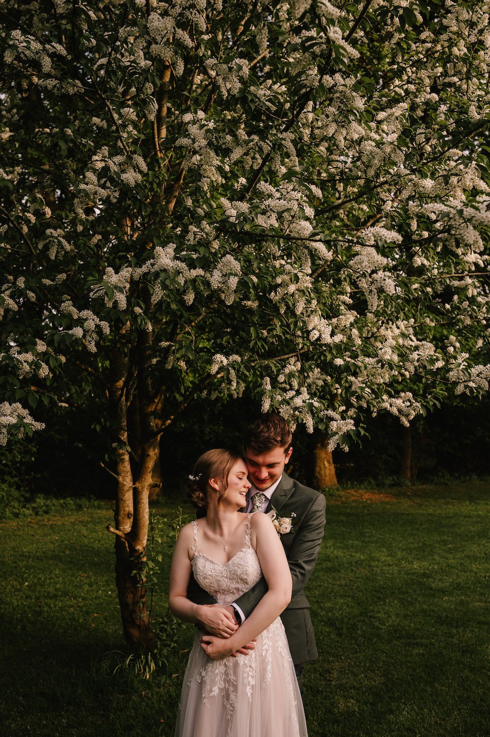 A bride and groom embrace beneath blooming trees in a park at sunset.