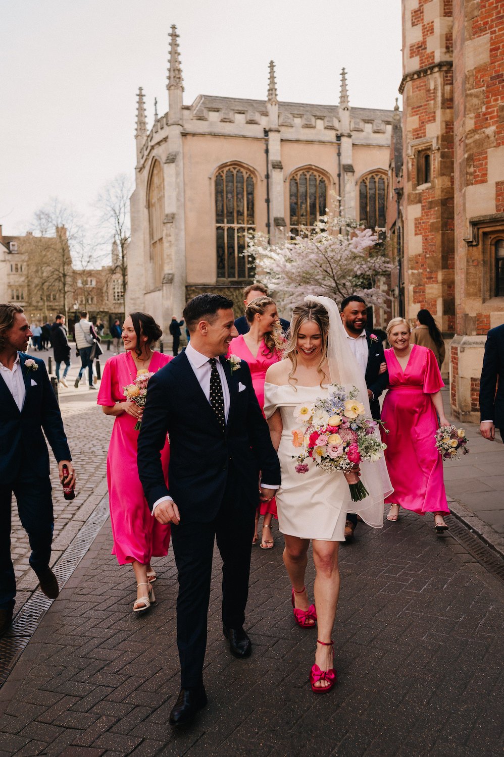 A wedding procession with the bride in a white dress and veil, holding a bouquet, walking with a groom in a dark suit. They are surrounded by bridesmaids in bright pink dresses and groomsmen in dark suits, all smiling and walking outdoors near histor