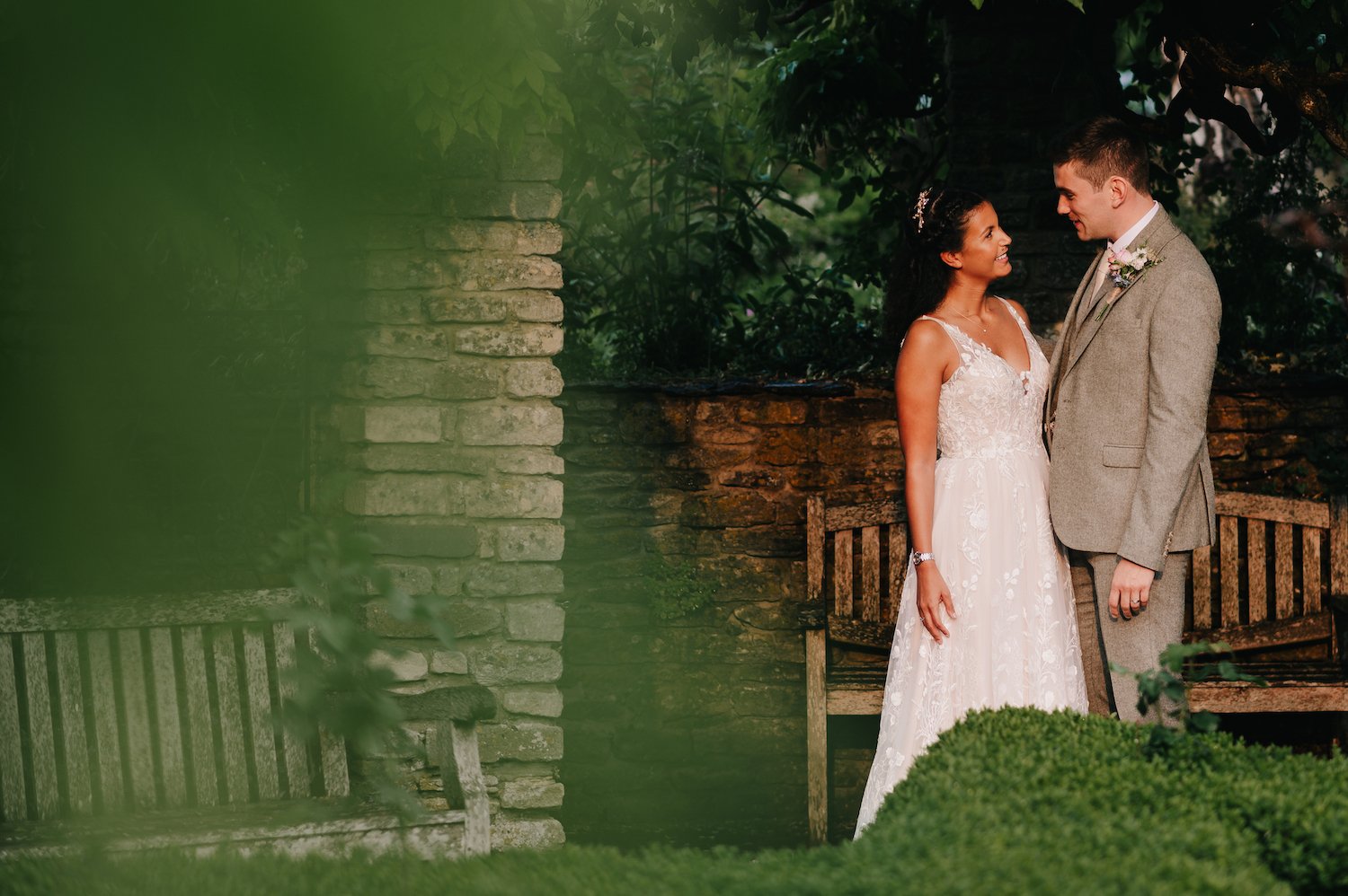 A bride and groom standing face to face outdoors, surrounded by greenery and a stone wall, sharing a tender moment on their wedding day.