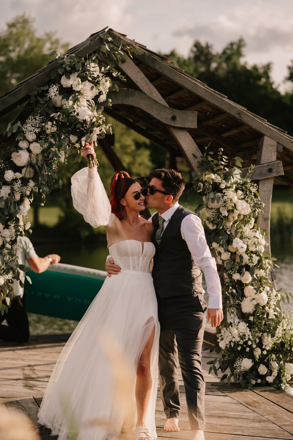 A bride and groom kiss during their outdoor wedding ceremony at the Jetty in Bedford, under a floral arch with white roses and greenery.