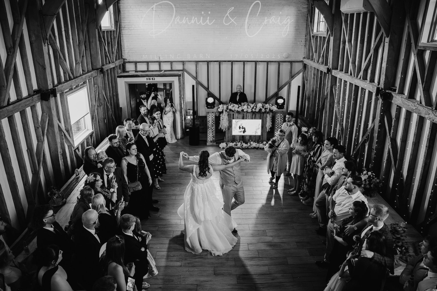 A black and white photo of a wedding reception with a bride and groom dancing in the center, surrounded by guests watching and smiling. The venue has rustic wooden walls and a decorative table at the front.