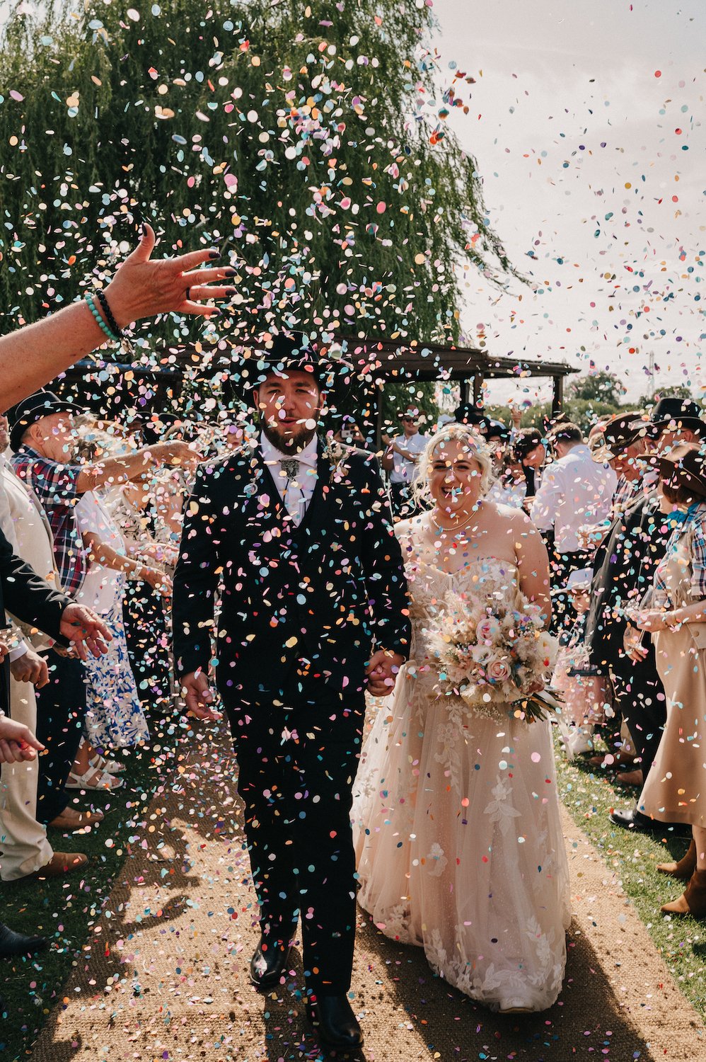 A newlywed couple holding hands walks through celebrating guests throwing colorful confetti at an outdoor wedding reception.